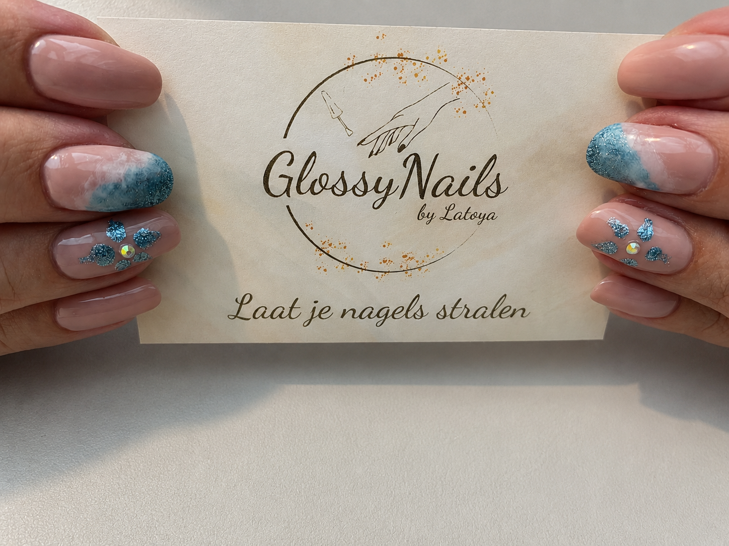 Handen met nude en blauwe nagels houden het kaartje van Glossy Nails by Latoya vast